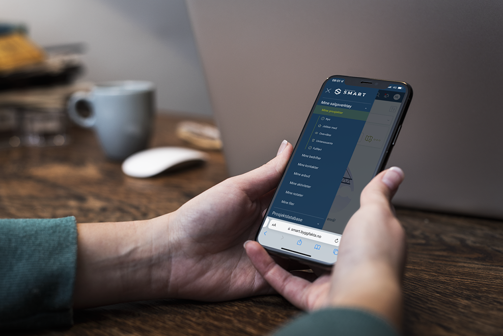 En person holder en mobiltelefon som viser prosjektdatabasen SMART. Skjermen viser en oversikt over byggeprosjekter med detaljerte data. Personen ser ut til å navigere i appen, som er tydelig synlig på telefonens skjerm.